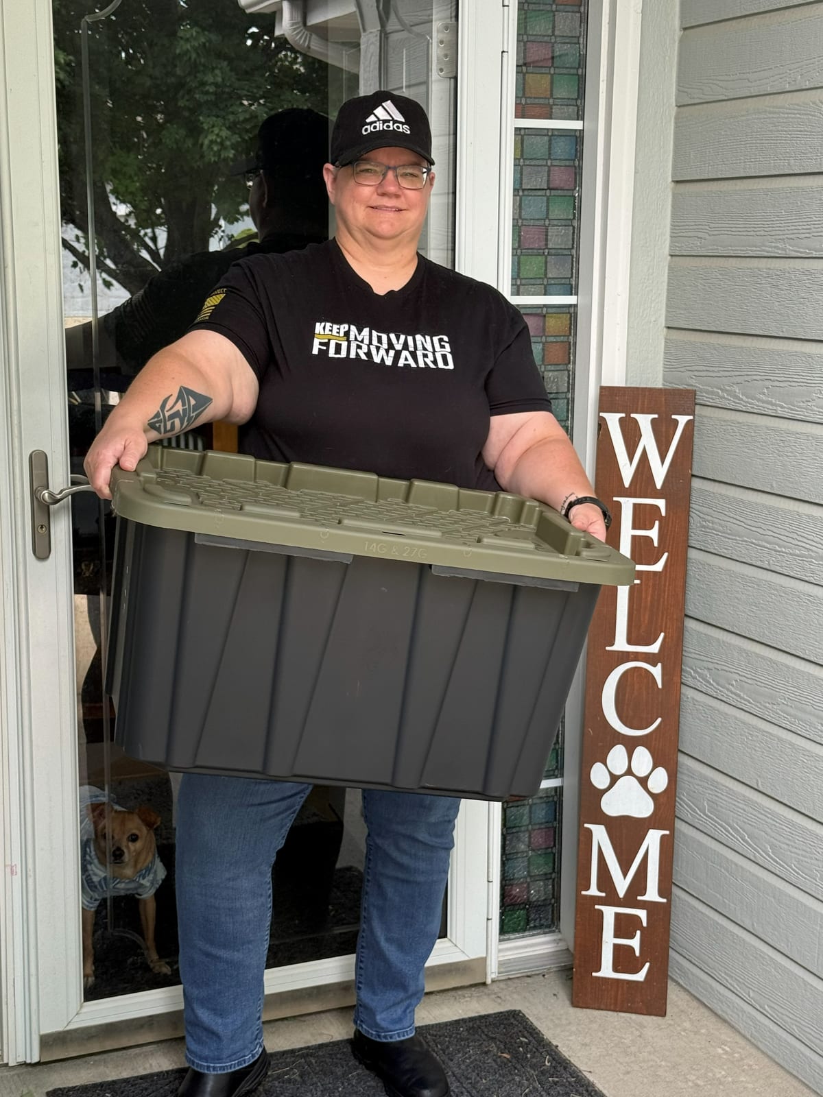 Shelley Fife standing at a front door holding a tote with a green lid, next to a welcome sign